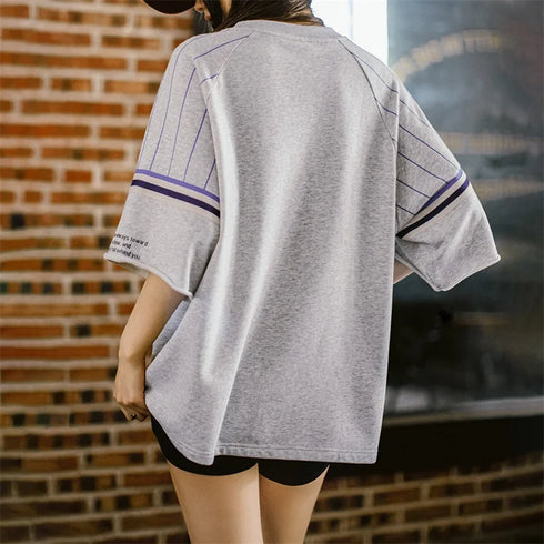 Person wearing a gray oversized t-shirt with purple accents in front of a brick wall.